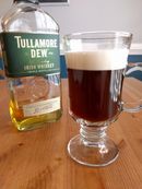 An Irish Coffee in a decorative glass mug. It has a thick layer of cream floating atop deep brown coffee. A bottle of Tullamore Dew Irish Whiskey is behind the mug.