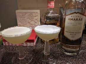 Two white cocktails with a frothy top in front of a bottle of mezcal and a bottle of tequila