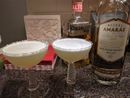 Two white cocktails with a frothy top in front of a bottle of mezcal and a bottle of tequila