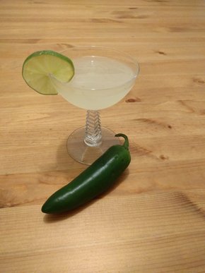 Cocktail in a coupe glass, garnished with a lime wheel. A jalapeño is on the table in front of it.