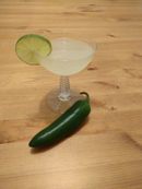 Cocktail in a coupe glass, garnished with a lime wheel. A jalapeño is on the table in front of it.